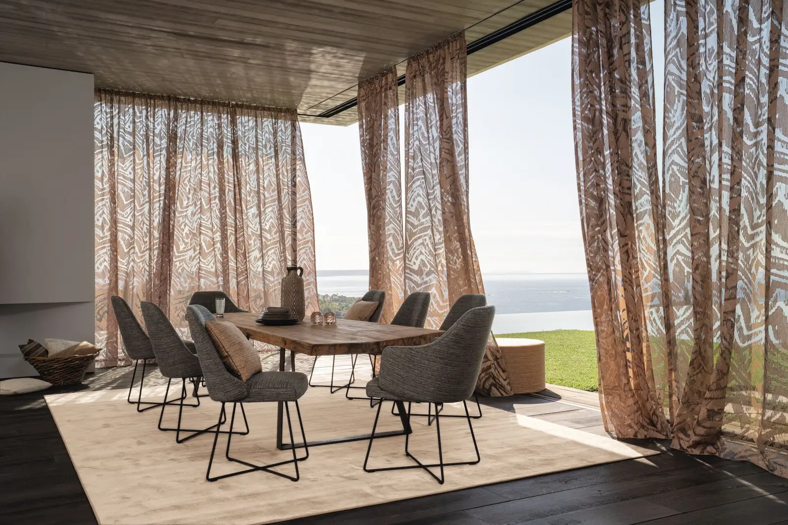 Transparente gemusterte Vorhänge bewegen sich im Wind vor einer offenen Terrasse mit Blick aufs Meer