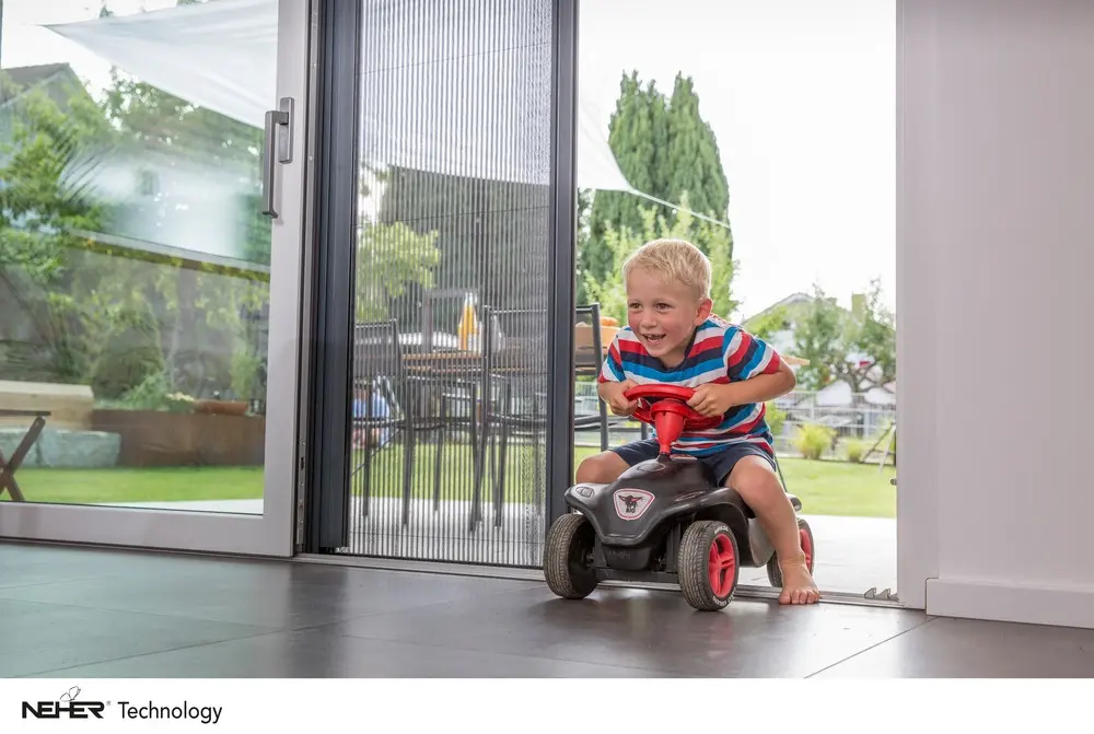 Kind fährt auf einem Rutschauto durch eine offene Terrassentür mit Plissee-Insektenschutz