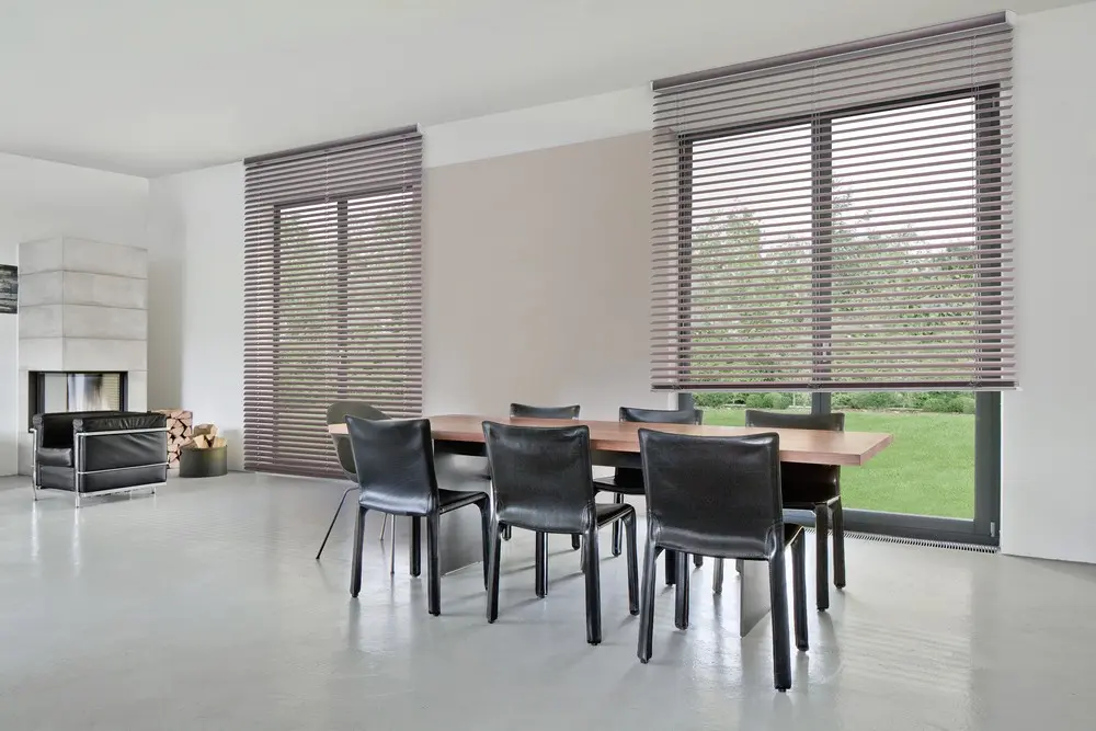 Zwei bodentiefe Fenster mit taupefarbenen Jalousien hinter einem Esstisch mit schwarzen Lederstühlen